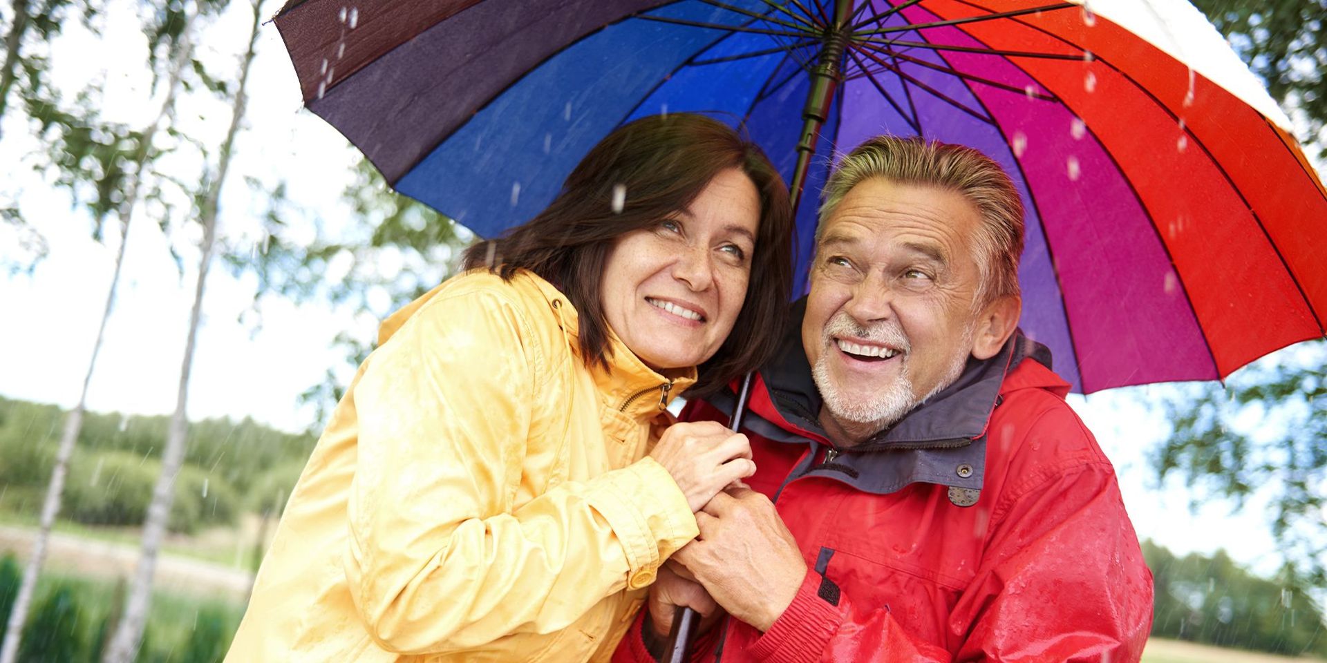 Älteres Paar lachend in Regenjacken unter einem bunten großen Regenschirm im Regen.